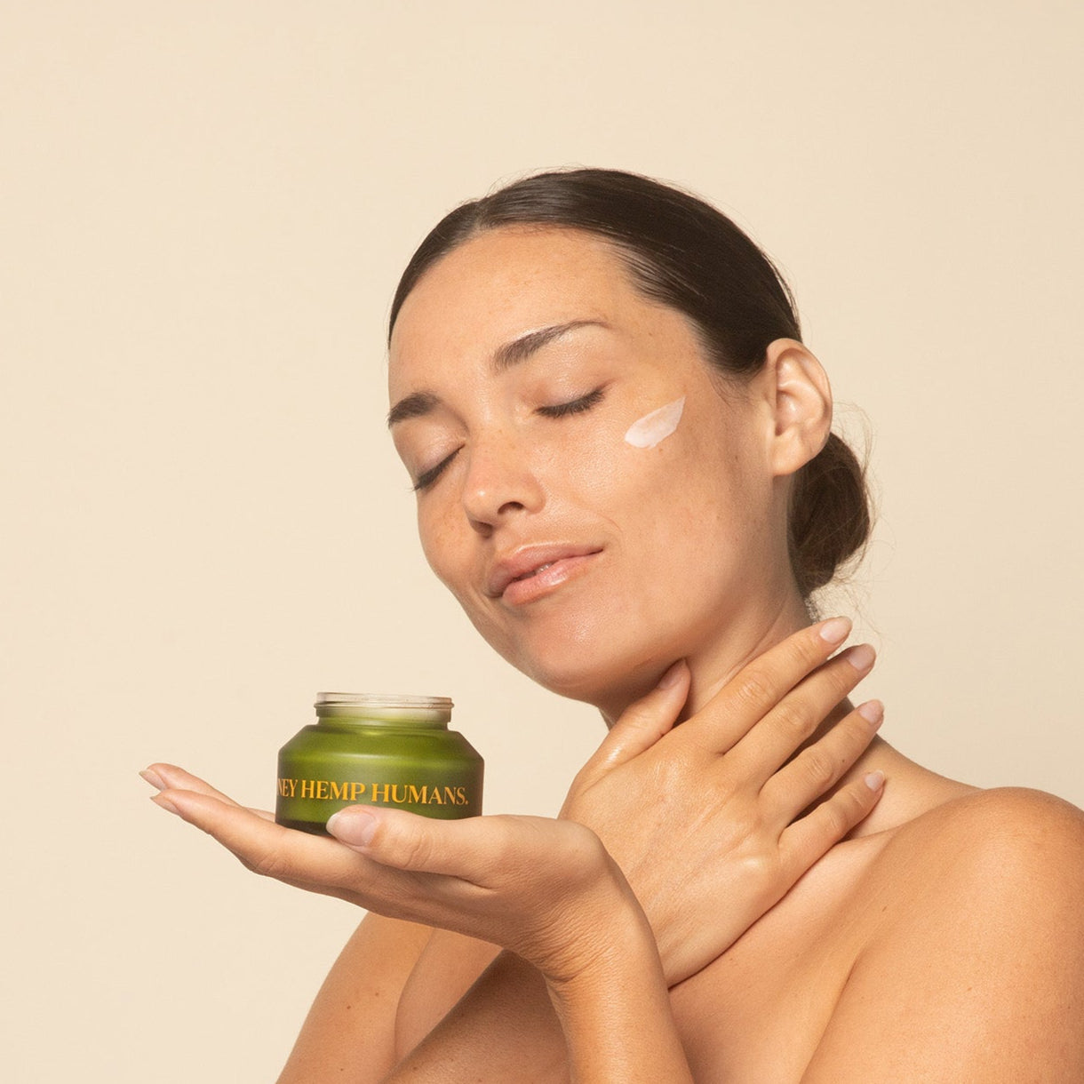 Woman applying apo ge manuka honey skincare-deep-hydration-face-cream to her neck with a jar of cream in her hand against a beige background