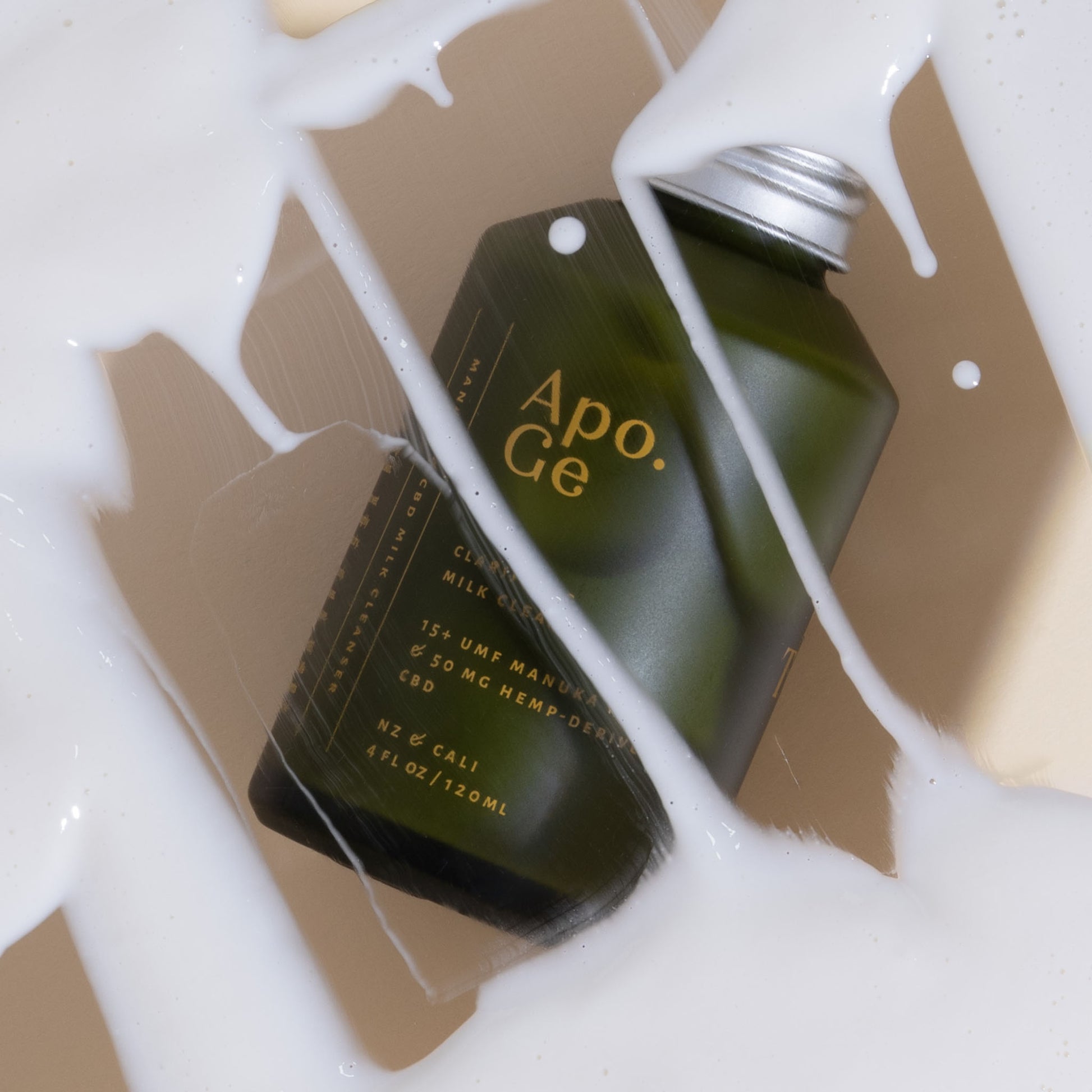 120ml green glass bottle with 'Apo.Ge' branding on a white background and apo ge manuka honey skincare clarifying milk cleanse spilled on background 