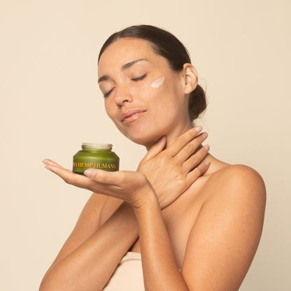 Woman applying cream to her face with a green jar labeled 'Honey Hemp Humans' on a beige background